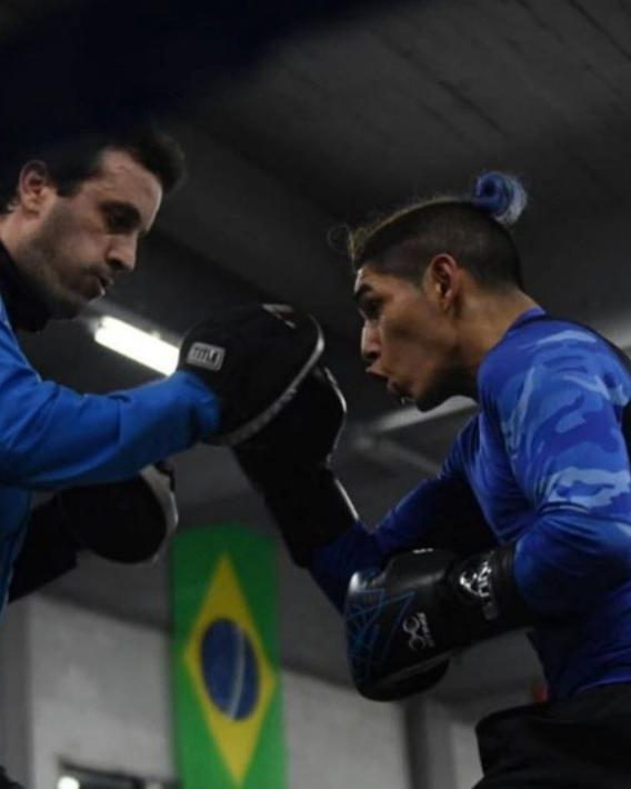 Calabrese sostiene las manoplas frente a un boxeador de remera azul que lanza un golpe, en un gimnasio con una bandera de Brasil al fondo