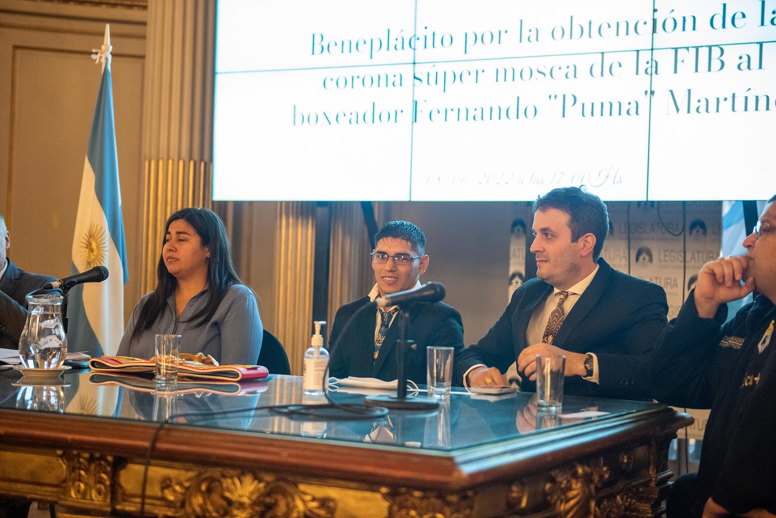 Calabrese y Fernando «Puma» Martínez en la mesa del acto de la Legislatura porteña; en la pantalla se lee «Beneplácito por la obtención de la corona súper mosca de la FIB al boxeador Fernando “Puma” Martínez» y la fecha 08/06/2022